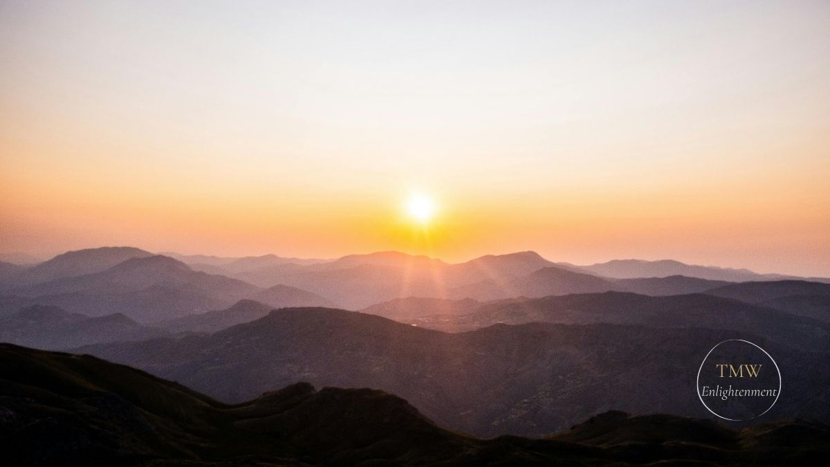 A rising round, orange sun just above the rolling hills and mountains.
